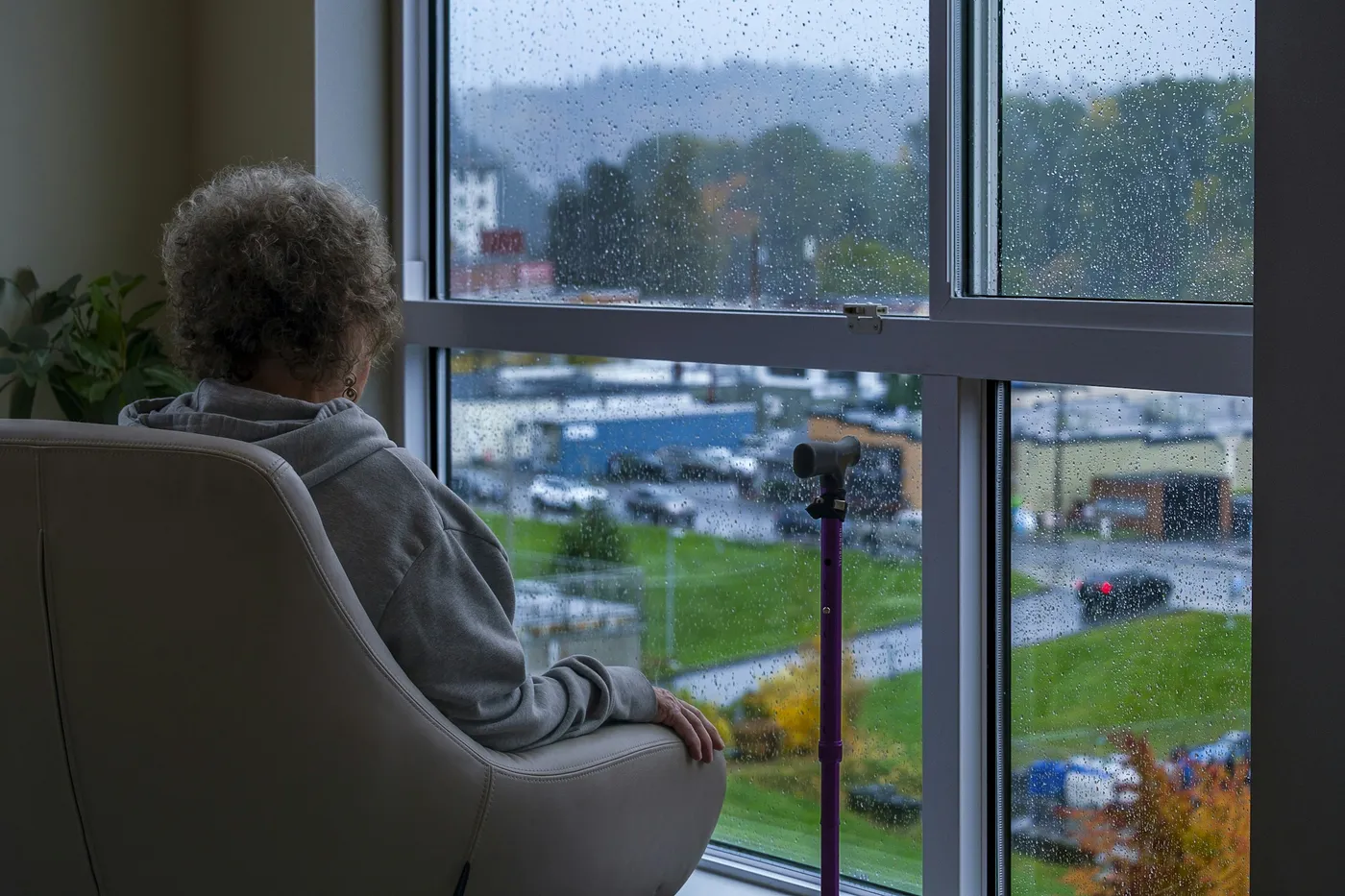 An older woman with a walking stick sits in an armchair, looking out of a rainy window.